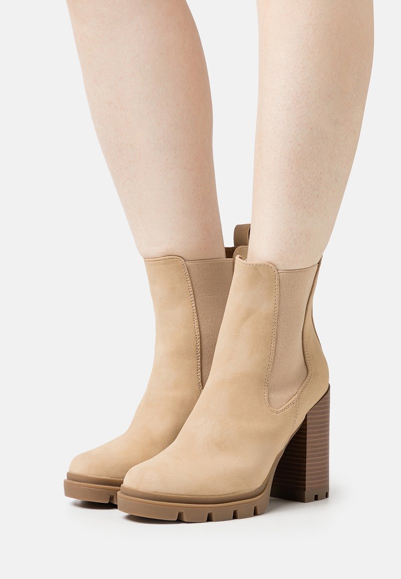 Bottes beiges en daim jusqu'à la cheville avec talons hauts en bois massif et panneaux latéraux élastiques, portées sur des jambes nues.