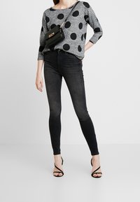 Top gris de manga larga con lunares negros, combinado con jeans ajustados negros y tacones negros de tiras. Incluye un pequeño bolso cruzado negro.