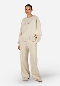 Bežs flīsa džemperis un platās bikses ar ribinātām piedurknēm, abiem apģērba gabaliem ir neliels melns "PEGADAS" uzraksts priekšpusē.