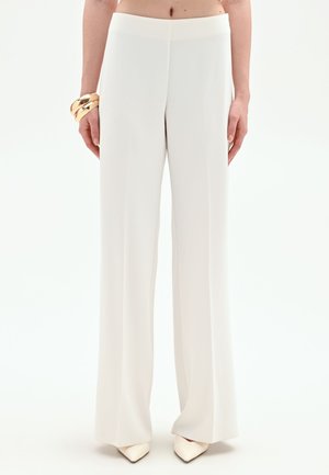 Donna che indossa pantaloni bianchi a vita alta e gamba larga, scarpe beige con punta e braccialetti dorati al polso sinistro, in piedi davanti a uno sfondo bianco.