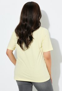 Jasnożółta koszulka z krótkim rękawem o luźnym kroju i prostym dole, noszona z jasnoszarymi spodniami. Włosy falowane, sięgające ramion.