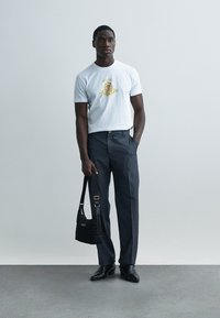 Weißes kurzärmliges T-Shirt mit goldenem Logo, kombiniert mit dunkler, maßgeschneiderter Hose. Schwarze gesteppte Schultertasche mit goldenen Details. Schwarze Schuhe.