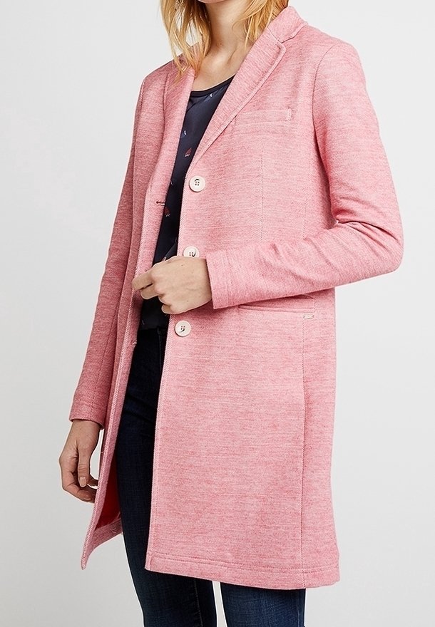 Femme portant un manteau rose boutonné longueur genou sur un haut noir et un jean bleu, debout devant un fond clair uni.