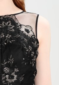 Černé šaty bez rukávů s průsvitnou vrstvou, zdobené propracovanou stříbrnou květinovou výšivkou a texturovanou látkou na živůtku.