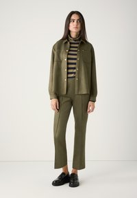 Olive green shirt jacket with gold buttons, striped turtleneck in olive and navy, tailored olive green pants, and black loafers.