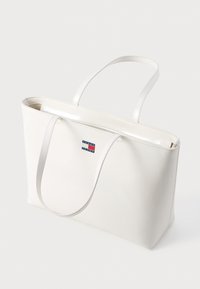 Geantă tote sintetică albă, cu textură netedă, mânere duble și o mică etichetă rectangulară cu logo în roșu, albastru și alb.