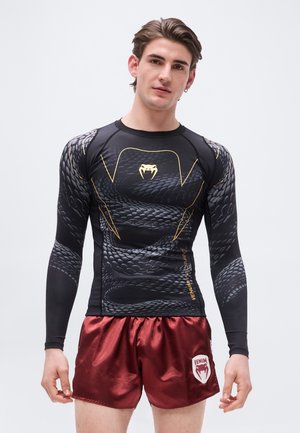 Młody mężczyzna ubrany w czarną kompresyjną bluzę z motywem węża oraz błyszczące bordowe spodenki z logo Venum stoi na tle jednolitego tła.