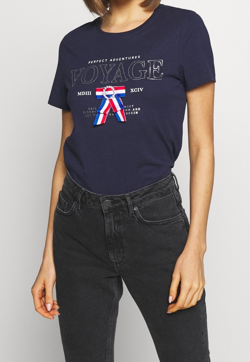T-shirt marine en coton, avec une impression graphique affichant le texte "VOYAGE" et un motif de ruban en rouge, blanc et bleu. Manches courtes.