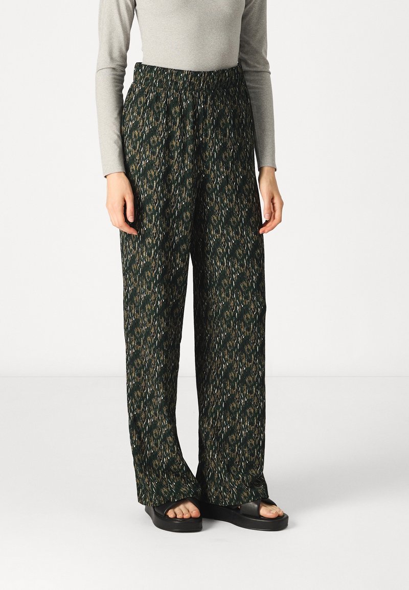 Personne portant un pantalon ample, taille haute, sombre à motifs, un haut gris à manches longues et des sandales compensées noires à bout ouvert.