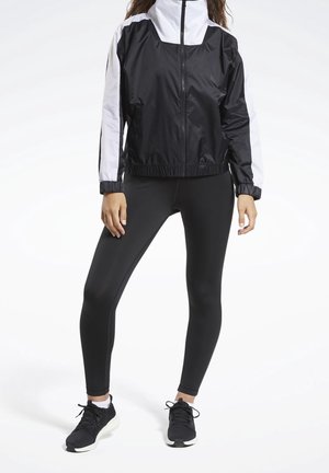 Mujer con chaqueta negra y blanca con cremallera, leggings negros y zapatillas negras de correr, de pie sobre fondo blanco.