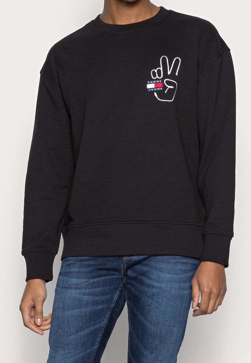 Persona che indossa una felpa nera con un simbolo della pace bianco e il logo Tommy Jeans sul petto, abbinata a jeans blu.
