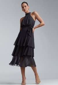 Musta halterneck-mekko, tehty virtaavasta materiaalista, jossa on kerroksittain laskostettuja röyhelöitä ja solmittava vyö vyötäröllä. Sileä pinta, polven mittainen helma.