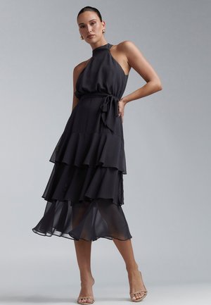 Melnas krāsas haltera kakla kleita no plūstoša materiāla ar kārtainiem volāniem un jostu viduklī. Gluda tekstūra, kleitas garums līdz ceļiem.