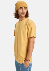 Žluté bavlněné tričko s krátkým rukávem a volným střihem, se malým logem na hrudi. Slučuje se s odpovídající pletenou žlutou čepicí.