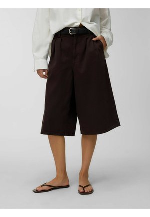 Personne portant un short ample foncé jusqu'aux genoux, une chemise blanche à manches longues, une ceinture noire et des sandales marron style tongs, debout sur un sol gris.
