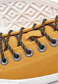 Žluté plátěné tenisky s texturovaným povrchem, černými tkaničkami a šedými očky, s bílou gumovou podrážkou se geometrickým vzorem.