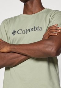 Světlé zelené tričko s krátkým rukávem a kulatým výstřihem, s tmavým logem. Tkanina se zdá být hladká a těsně obepíná tělo.