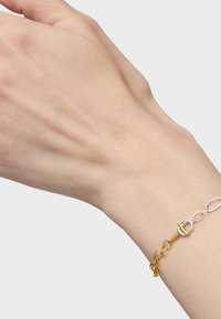 Braccialetto a catena in oro e argento su un polso, caratterizzato da maglie intrecciate e una chiusura decorativa con forme e finiture distintive.