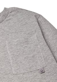 Hellgraues Strickshirt, das eine Nahaufnahme einer Brusttasche zeigt, mit einem kleinen Markenschild am unteren Rand.
