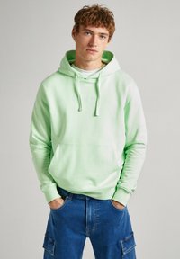 Sudadera verde menta con cordones y bolsillo frontal, con bordado de logotipo sutil, combinada con jeans azules y una camisa blanca.