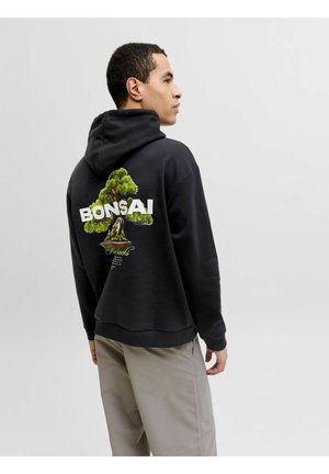 Mężczyzna w czarnej bluzie z kapturem z grafiką drzewa bonsai i napisem na plecach, beżowe spodnie, patrzący na bok na jednolitym tle.