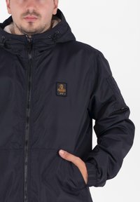 Giacca con cappuccio blu navy, dotata di zip frontale, foderata in pile e con logo ricamato sul petto. Presenta polsini elasticizzati e tasche laterali.