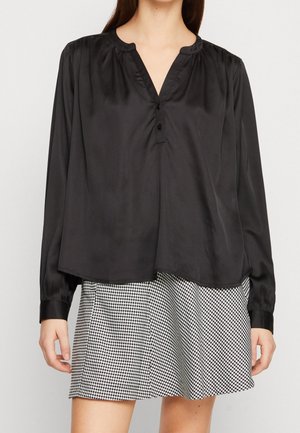 Vrouw draagt een losse zwarte satijnen blouse met lange mouwen en een minirok met pied-de-poule motief, gefotografeerd tegen een effen achtergrond.