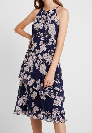Femme portant une robe bleu marine sans manches avec un imprimé floral blanc et un ourlet à volants superposés, debout avec une main sur la hanche.