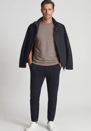 Man draagt een taupekleurig T-shirt, zwarte broek, zwarte jas over de schouders gedrapeerd, met handen in de zakken en witte sneakers, staand tegen een effen achtergrond.