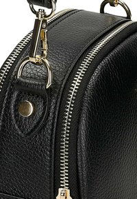 Primo piano di un angolo di una borsa in pelle nera texture con cerniera argentata, chiusura in metallo e tracolla in pelle cucita con rivetto.