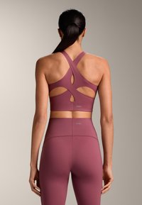 Donna con coda di cavallo scura che indossa un reggiseno sportivo malva con ritagli a incrocio e leggings coordinati a vita alta, in piedi davanti a uno sfondo semplice.