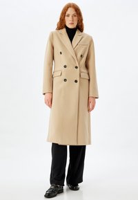 Cappotto beige doppiopetto con bottoni neri, tasche laterali e colletto a punte. Abbinato a un dolcevita nero e pantaloni neri.
