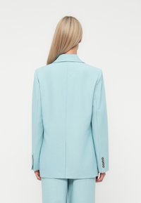 Woman with long blonde hair wearing a light blue tailored blazer and matching pants, shown from the back against a plain white background.