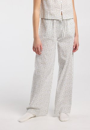 Femme portant un pantalon de pyjama ample blanc à motif floral et un haut sans manches assorti, debout pieds nus sur un fond uni.