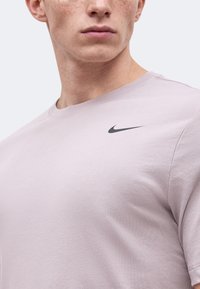 Νεαρός με ανοιχτόχρωμο δέρμα που φορά ένα ανοιχτό μωβ T-shirt Nike με μαύρο λογότυπο swoosh στο αριστερό στήθος.