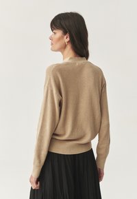 Femme aux cheveux foncés portant un pull en maille beige et une jupe plissée noire, se tenant de profil contre un fond clair.