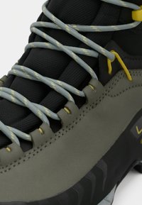 Stivali da trekking allacciabili in nero e verde oliva, con lacci grigi e dettagli gialli, materiale testurizzato e un design della suola robusto.