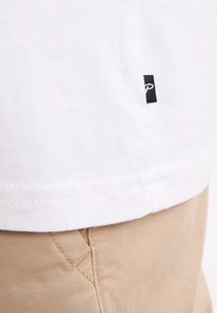 Camiseta blanca de algodón con una pequeña etiqueta negra en el dobladillo, combinada con pantalones de algodón beige claros que presentan una costura detallada.