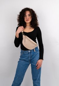 Beige Leder Umhängetasche mit geschwungener Form und goldener Reißverschluss. Das Modell trägt ein schwarzes Oberteil und blaue hochtaillierte Jeans und steht vor einem weißen Hintergrund.