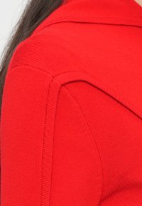 Îmbrăcăminte tricotată roșie cu o textură netedă, cu umeri structurați și detalii de cusătură, evidențiind o culoare vibrantă.