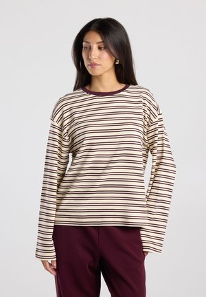 Woman wearing a cream and dark brown horizontal striped long-sleeve shirt and dark burgundy pants, looking to the side.