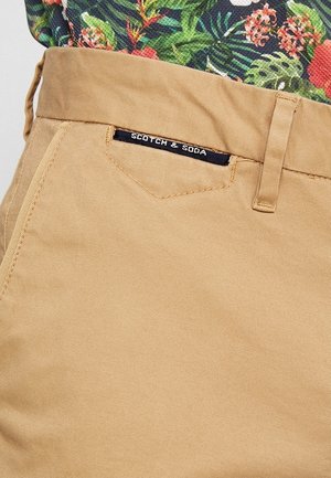Kieszeni w spodniach w kolorze jasnobeżowym z czarną metką z napisem "Scotch & Soda" oraz fragment zielono-czerwonej kwiecistej koszuli nad pasem.