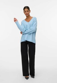 Femme se tenant debout, portant un pull V-neck bleu clair, un pantalon large noir et des chaussures à talons noires à pois blancs, posant sur un fond blanc.