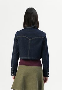 Giacca di jeans con un design cropped, colore blu scuro e polsini con bottoni, indossata su un maglione bordeaux e una gonna verde oliva.
