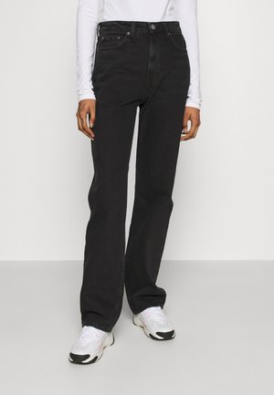 Personne portant un jean noir taille haute à jambes droites, un haut blanc à manches longues et des baskets blanches, debout devant un fond uni.