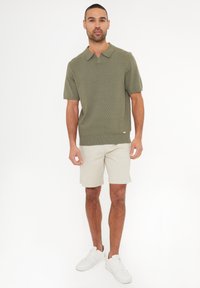 Kortærmet olivengrøn polo t-shirt med et struktureret zigzag-mønster, parret med lysebeige shorts og hvide sneakers.