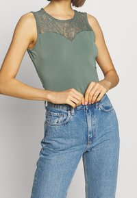 Ärmlös topp i dämpad grön tyg med spetsdetaljer runt halsringningen, matchad med ljusblå högmidjade denimjeans.