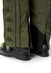 Pantaloni verde oliva con strisce laterali con logo indossati sopra scarponi da sci neri e verde neon, mostrando le gambe posteriori e i polpacci inferiori.