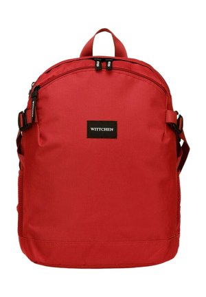 Mochila de lona roja con forma redondeada, compartimento frontal con cremallera, bolsillo de malla lateral y un parche de logo negro. Incluye correas ajustables.