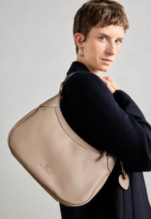 Beige Leder Schultertasche mit strukturiertem Finish, geschwungener Form und einem goldenen Akzentanhänger, der an dem Riemen befestigt ist.